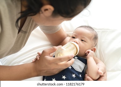 Young Hispanic Baby Or Asian Infant Boy Drinking Milk From Plastic Bottle Feeding From Young Parents Mother Or Babysitter With Love And Bonding. Child Care From Mom Holding Kid In Arm On Blanket Bed.