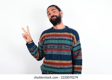 young hispanic bearded man wearing knitted sweater over white background makes peace gesture keeps lips folded shows v sign. Body language concept