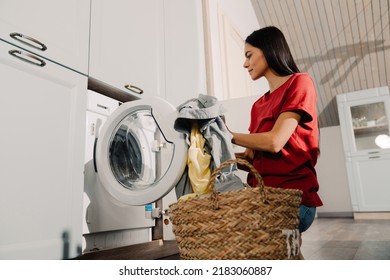 Young hispanic brunette woman putting clothes at washing machine while doing laundry at home