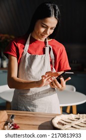 Young Hispanic Brunette Woman Wearing Apron Taking Photo Of Meal While Cooking In Kitchen At Home