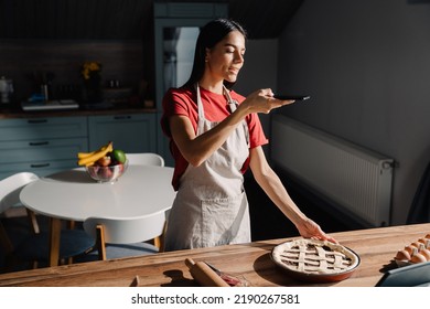 Young Hispanic Brunette Woman Wearing Apron Taking Photo Of Meal While Cooking In Kitchen At Home