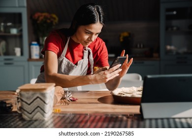 Young Hispanic Brunette Woman Wearing Apron Taking Photo Of Meal While Cooking In Kitchen At Home