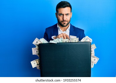 Young Hispanic Businessman Holding Briefcase Full Of Dollars Skeptic And Nervous, Frowning Upset Because Of Problem. Negative Person. 