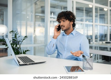 Young Hispanic businessman looking frustrated and angry during a phone call in a modern office setting with laptop and tablet on desk. - Powered by Shutterstock