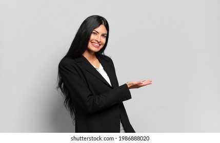 young hispanic businesswoman smiling cheerfully, feeling happy and showing a concept in copy space with palm of hand