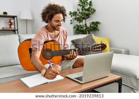 Young hispanic composer man composing song using guitar and laptop at home.