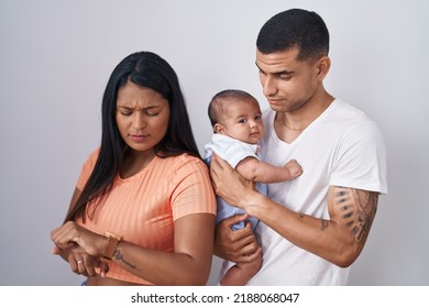 Young Hispanic Couple With Baby Standing Together Over Isolated Background Checking The Time On Wrist Watch, Relaxed And Confident 