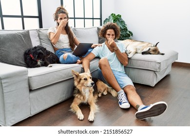 Young hispanic couple with dogs relaxing at home smelling something stinky and disgusting, intolerable smell, holding breath with fingers on nose. bad smell 
