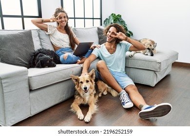 Young hispanic couple with dogs relaxing at home doing peace symbol with fingers over face, smiling cheerful showing victory 