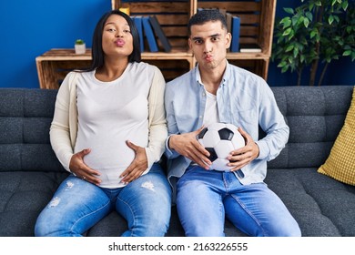Young hispanic couple expecting a baby sitting on the sofa holding ball and tummy looking at the camera blowing a kiss being lovely and sexy. love expression. 