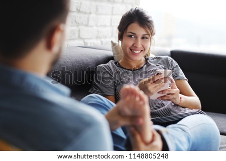 Young hispanic couple laying on couch at home, using a mobile phone for Internet and social media. The boyfriend is giving a massage to feet of the woman.