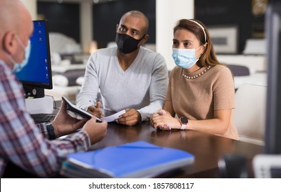 Young Hispanic Couple In Protective Masks Consulting With Salesman When Choosing New Mattress In Furniture Store. Concept Of Shopping Precautions And Social Distancing In Pandemic