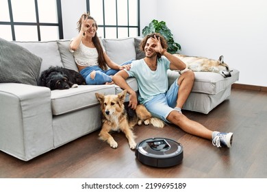 Young hispanic couple relaxing at home by vacuum robot smiling happy doing ok sign with hand on eye looking through fingers 