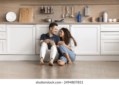 Young Hispanic couple resting seated on warm floor in fashionable kitchen, enjoy pleasant conversation, drinking beverage, spend relocation day to their first own house together. Cohabitation, love - Powered by Shutterstock