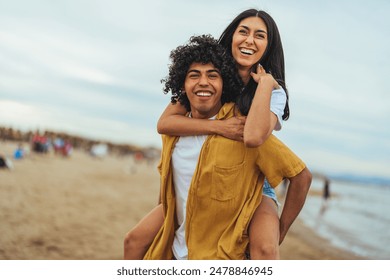 A young Hispanic couple shares a playful moment with a piggyback ride along the shore, dressed in casual summer clothes, embodying the carefree spirit of a beach outing. - Powered by Shutterstock