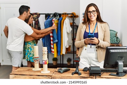 Young Hispanic Couple Shopping At Clothes Store. Shopkeeper Woman Smiling Happy And Using Smartphone.