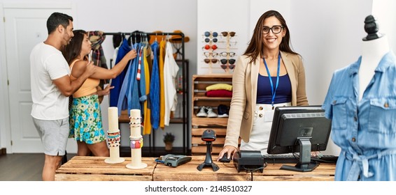 Young Hispanic Couple Shopping At Clothes Store. Shopkeeper Woman Smiling Happy Standing By Fashion Shop Counter.