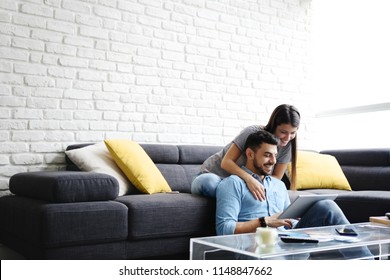 Young hispanic couple sitting on couch at home, using a tablet PC for Internet and social media. The girl is giving a massage to her boyfriend. Copy space