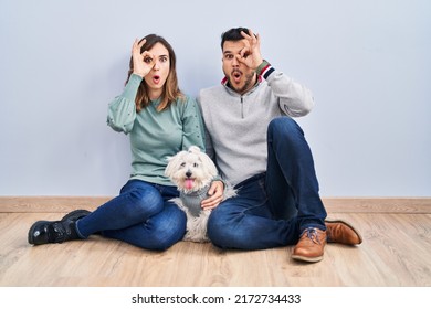 Young Hispanic Couple Sitting On The Floor With Dog Doing Ok Gesture Shocked With Surprised Face, Eye Looking Through Fingers. Unbelieving Expression. 