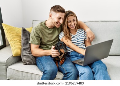 Young Hispanic Couple Smiling Happy Using Laptop Sitting On The Sofa  With Dog At Home.