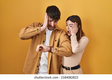 Young Hispanic Couple Standing Over Yellow Background Looking At The Watch Time Worried, Afraid Of Getting Late 