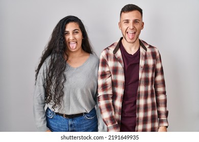 Young Hispanic Couple Standing Over White Background Sticking Tongue Out Happy With Funny Expression. Emotion Concept. 