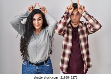 Young Hispanic Couple Standing Over White Background Doing Funny Gesture With Finger Over Head As Bull Horns 