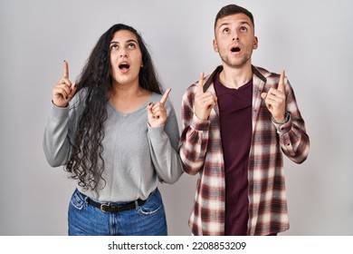 Young Hispanic Couple Standing Over White Background Amazed And Surprised Looking Up And Pointing With Fingers And Raised Arms. 