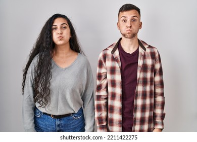 Young Hispanic Couple Standing Over White Background Puffing Cheeks With Funny Face. Mouth Inflated With Air, Crazy Expression. 