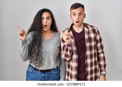 Young Hispanic Couple Standing Over White Background Surprised Pointing With Finger To The Side, Open Mouth Amazed Expression. 