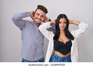 Young Hispanic Couple Standing Over White Background Relaxing And Stretching, Arms And Hands Behind Head And Neck Smiling Happy 