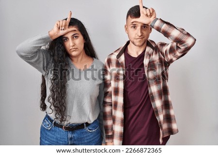 Young hispanic couple standing over white background making fun of people with fingers on forehead doing loser gesture mocking and insulting. 