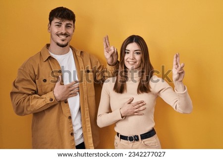 Young hispanic couple standing over yellow background smiling swearing with hand on chest and fingers up, making a loyalty promise oath 