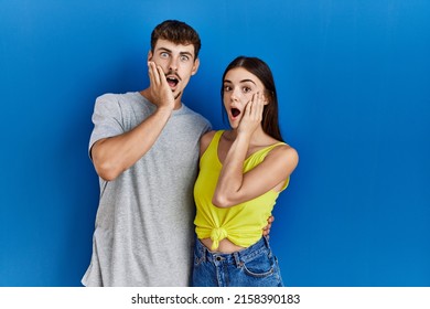 Young Hispanic Couple Standing Together Over Blue Background Afraid And Shocked, Surprise And Amazed Expression With Hands On Face 