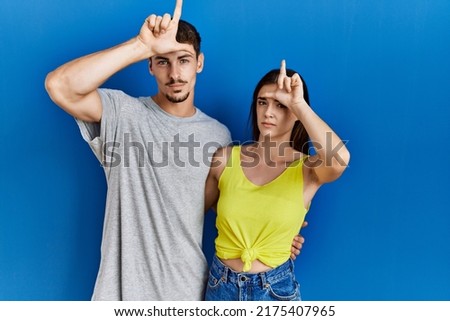 Young hispanic couple standing together over blue background making fun of people with fingers on forehead doing loser gesture mocking and insulting. 
