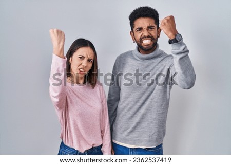 Young hispanic couple standing together angry and mad raising fist frustrated and furious while shouting with anger. rage and aggressive concept. 