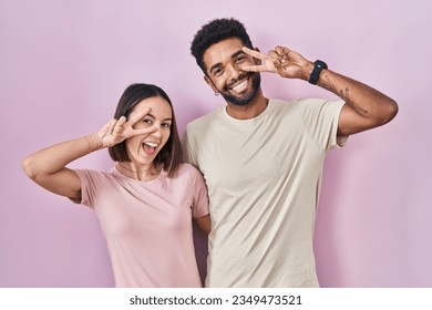 Young hispanic couple together over pink background doing peace symbol with fingers over face, smiling cheerful showing victory 
