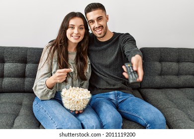 Young Hispanic Couple Watching Film And Eating Popcorn Sitting On The Sofa At Home.