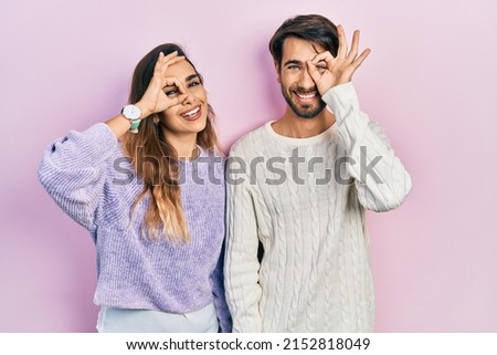 Young hispanic couple wearing casual clothes doing ok gesture with hand smiling, eye looking through fingers with happy face. 