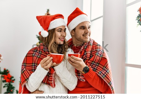 Young hispanic couple wearing christmas hat and covering with blanket. Standing with smile on face drinking cofffee at home.