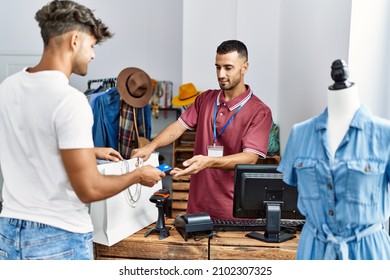 Young Hispanic Customer Man Paying To Shopkeeper Using Credit Card At Clothing Store.