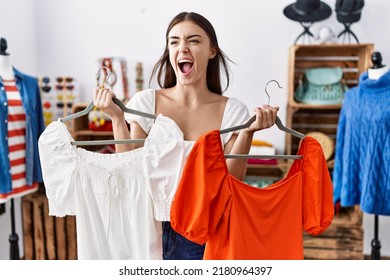 Young Hispanic Customer Woman Holding Shirts At Clothing Store Angry And Mad Screaming Frustrated And Furious, Shouting With Anger. Rage And Aggressive Concept. 