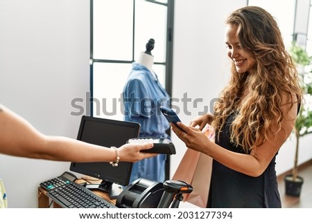 Young hispanic customer woman smiling happy paying using smartphone at clothing store.