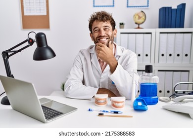 Young hispanic dentist man working at medical clinic looking confident at the camera smiling with crossed arms and hand raised on chin. thinking positive. 