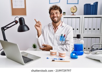 Young Hispanic Dentist Man Working At Medical Clinic With A Big Smile On Face, Pointing With Hand And Finger To The Side Looking At The Camera. 