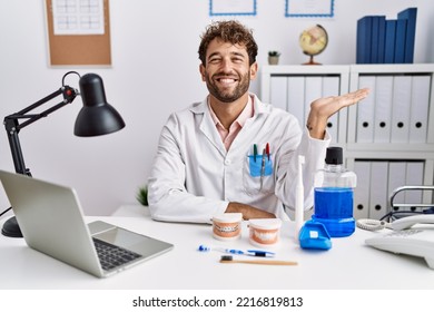 Young Hispanic Dentist Man Working At Medical Clinic Smiling Cheerful Presenting And Pointing With Palm Of Hand Looking At The Camera. 