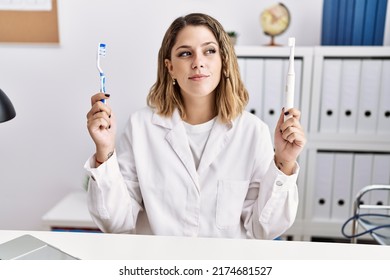 Young hispanic dentist woman holding electric toothbrush and teethbrush at clinic smiling looking to the side and staring away thinking. 