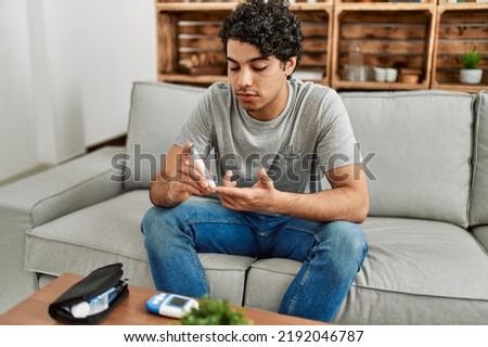 Young hispanic diabetic man measuring glucose sitting on the sofa at home.