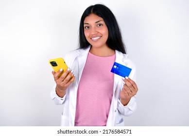 Young Hispanic Doctor Girl Wearing Coat Over White Background Holds Modern Mobile Phone And Credit Card