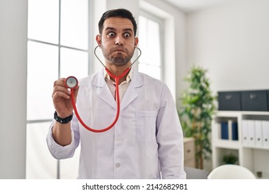 Young hispanic doctor man with beard holding stethoscope auscultating puffing cheeks with funny face. mouth inflated with air, catching air.  - Shutterstock ID 2142682981